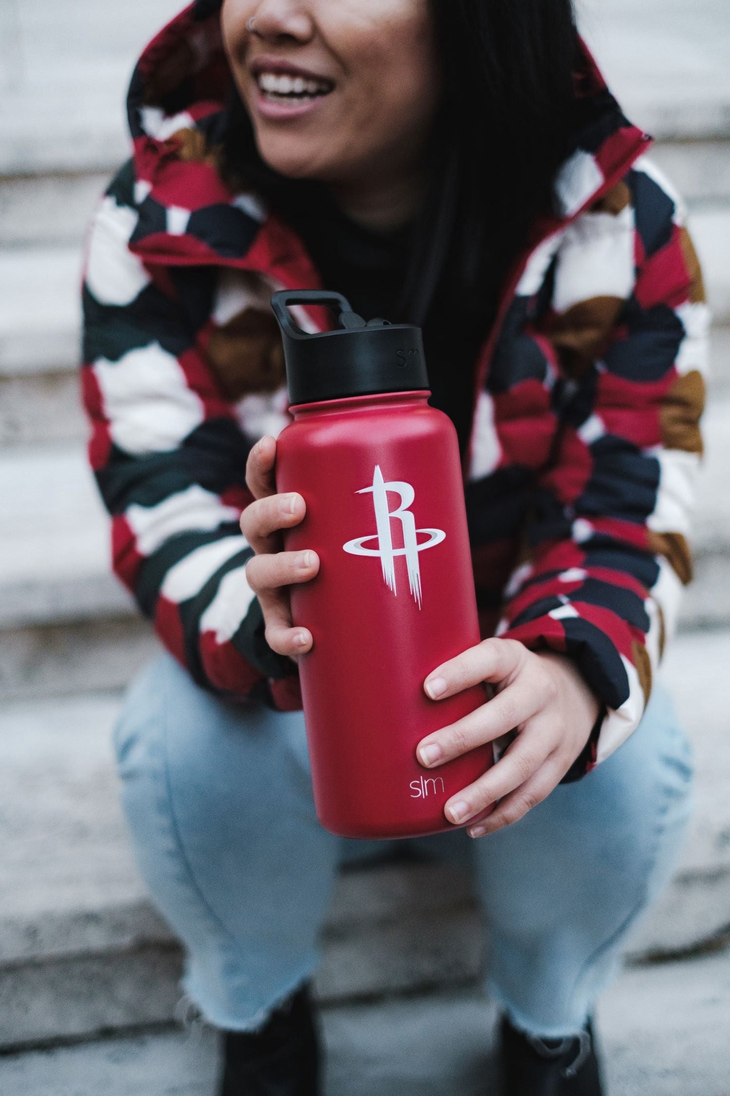 Red water bottle held in hand with person wearing color-blocked jacket in red, navy blue, white, and golden mustard tones