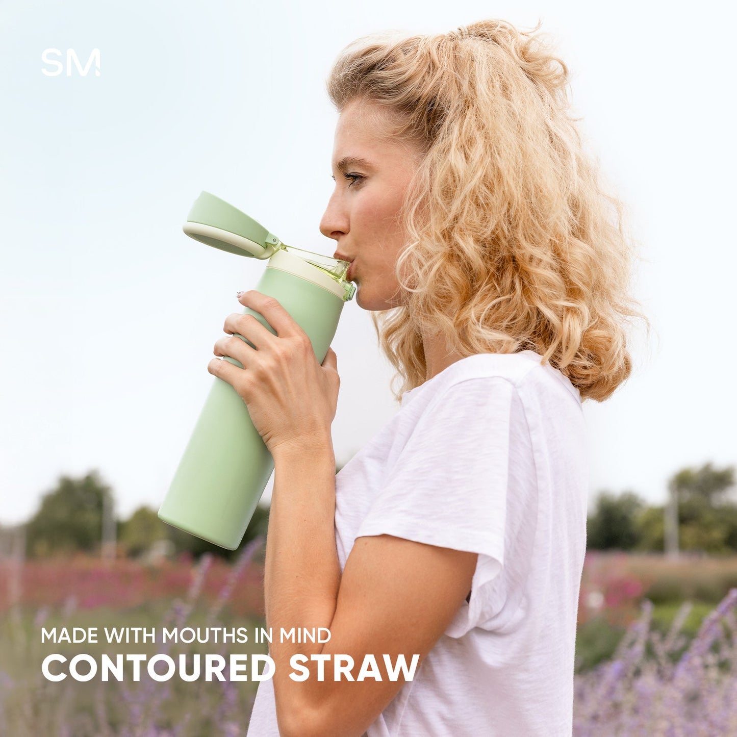 Woman with blonde hair wearing a pink shirt drinks from a sage green Mesa Loop cup with contoured straw in a lavender field. Simple Modern logo visible.