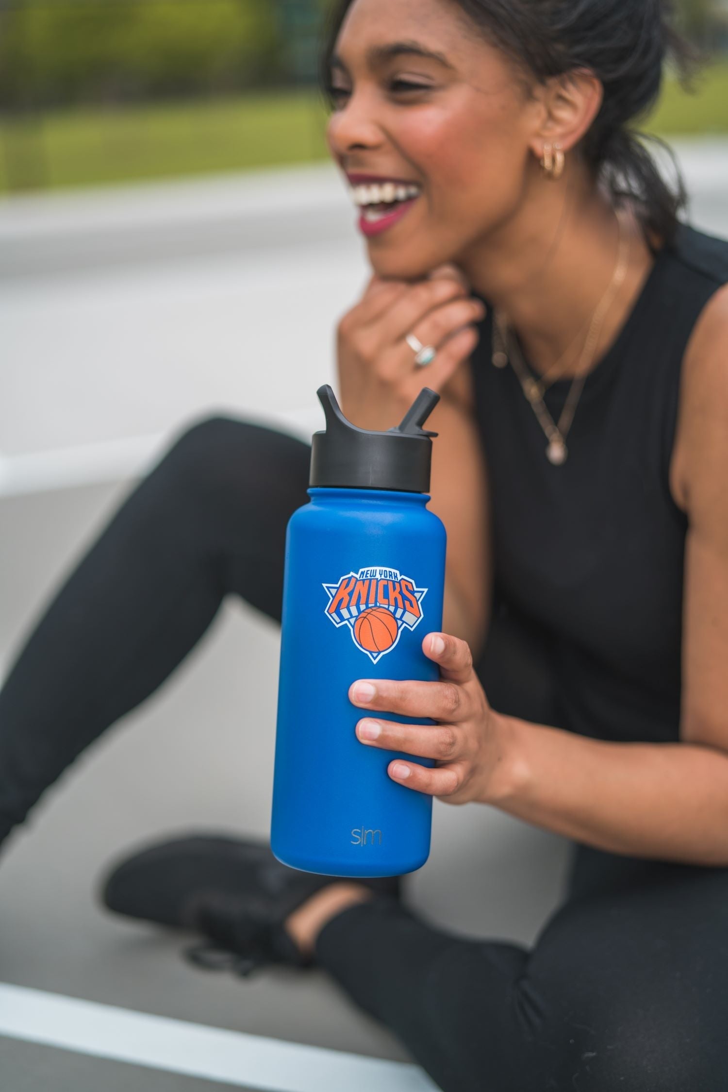 Royal blue water bottle held by person wearing black sleeveless top with green grass and neutral background