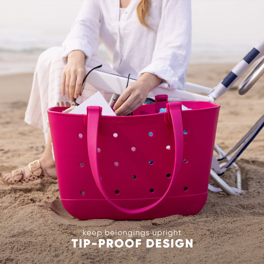 Hot pink tote bag with tip-proof design and drainage holes held by person in white shirt on beach. Text reads "keep belongings upright" and "TIP-PROOF DESIGN."
