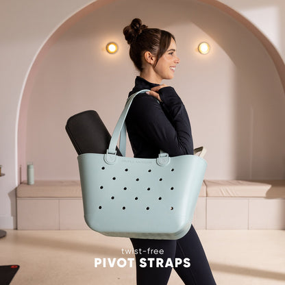 Woman in black athletic wear walking while carrying a pale blue-gray perforated tote bag with twist-free pivot straps, in a minimalist studio setting with warm lighting.