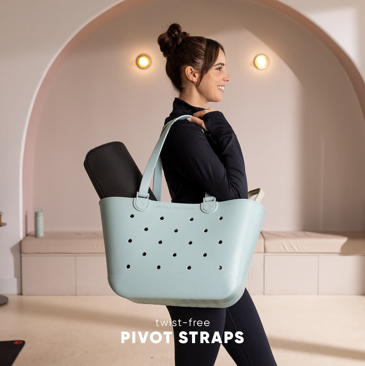 Woman in black athletic wear holding a light mint blue tote bag with black perforations and twist-free pivot straps in a modern studio with curved archway backdrop.
