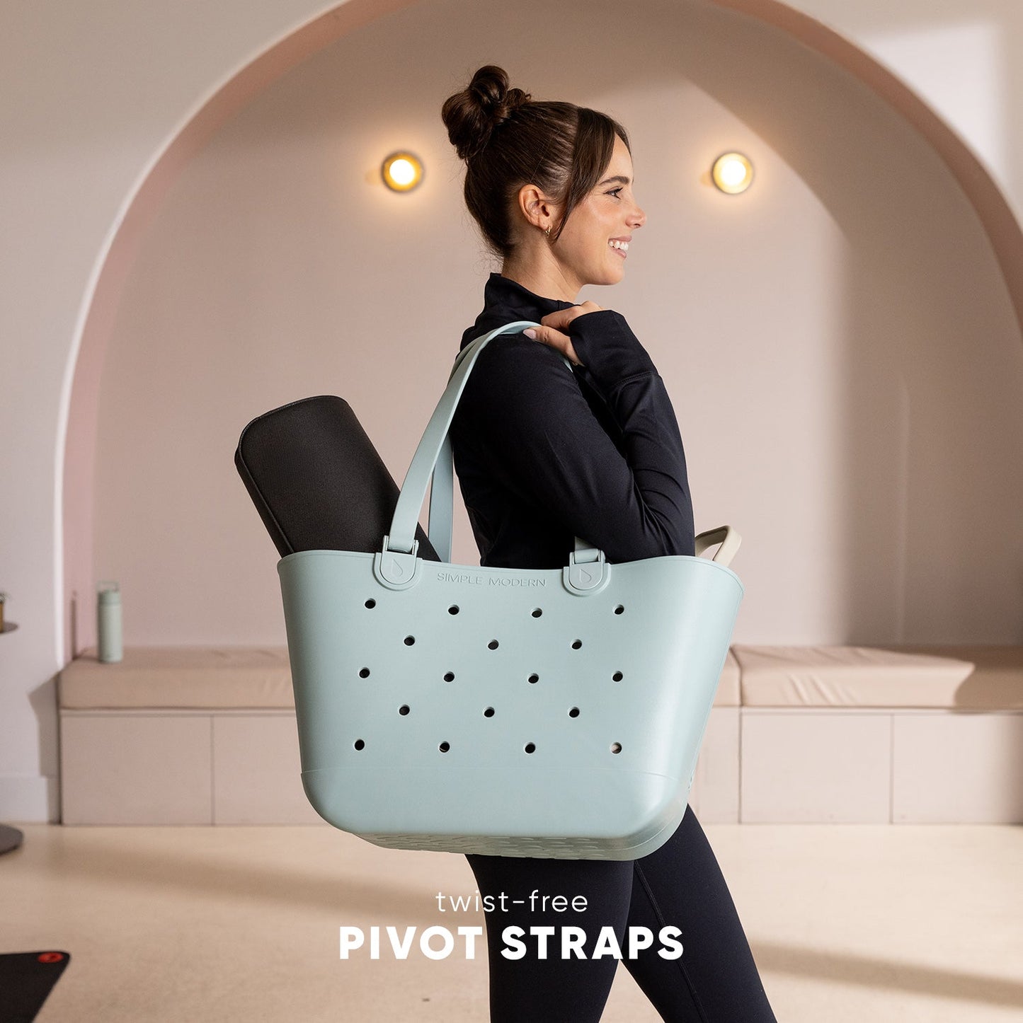 Woman wearing black athletic top and leggings carries a pale blue-green perforated tote bag with twist-free pivot straps over her shoulders in a modern studio setting with curved architecture.