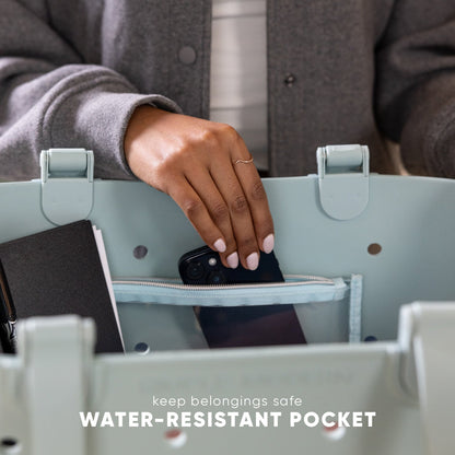 Person placing black smartphone into sage green tote bag's water-resistant pocket. Text reads "keep belongings safe WATER-RESISTANT POCKET." Lifestyle product demonstration showing bag functionality.