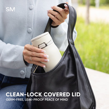 Cream Simple Modern tumbler with CLEAN-LOCK covered lid being placed into a black tote bag, held by a person wearing a light gray cardigan outdoors with blurred green background.