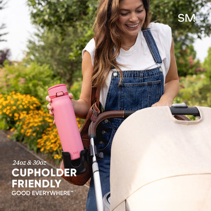 Woman in denim overalls holding a bright pink Simple Modern water bottle in a blooming garden with a cream stroller nearby. Text reads "24oz & 30oz Cupholder Friendly."