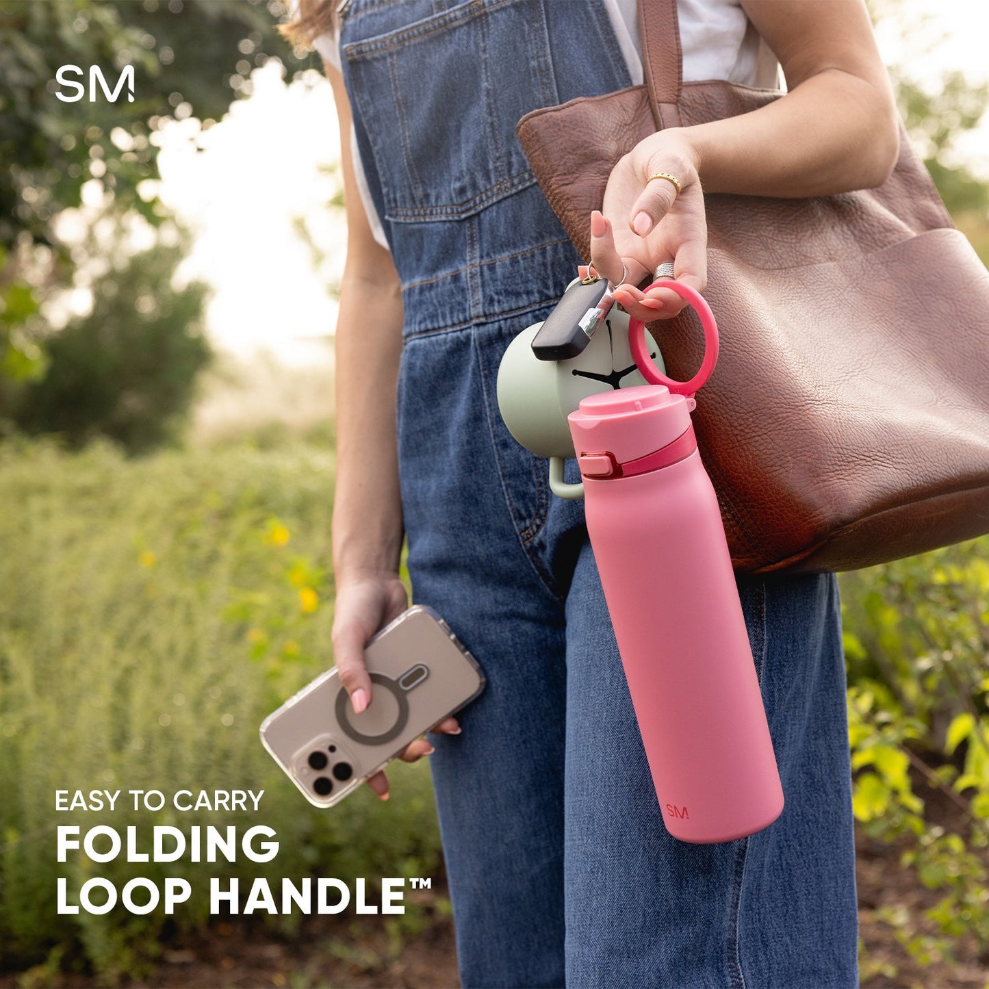 Person holding a bright pink Simple Modern water bottle with folding loop handle attached to a brown leather bag in an outdoor park setting. White text reads "Easy to Carry" and "Folding Loop Handle."