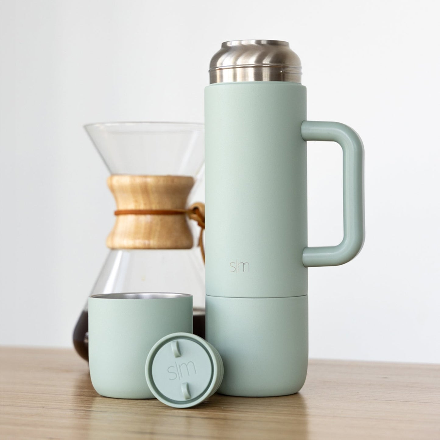 Sage green insulated tumbler and matching cup with warm wood and silver metallic accents on wooden surface