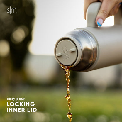 Metallic silver bottle with amber-golden liquid pouring out, set against a soft gray and white background with warm neutral tones and brushed stainless steel finish.