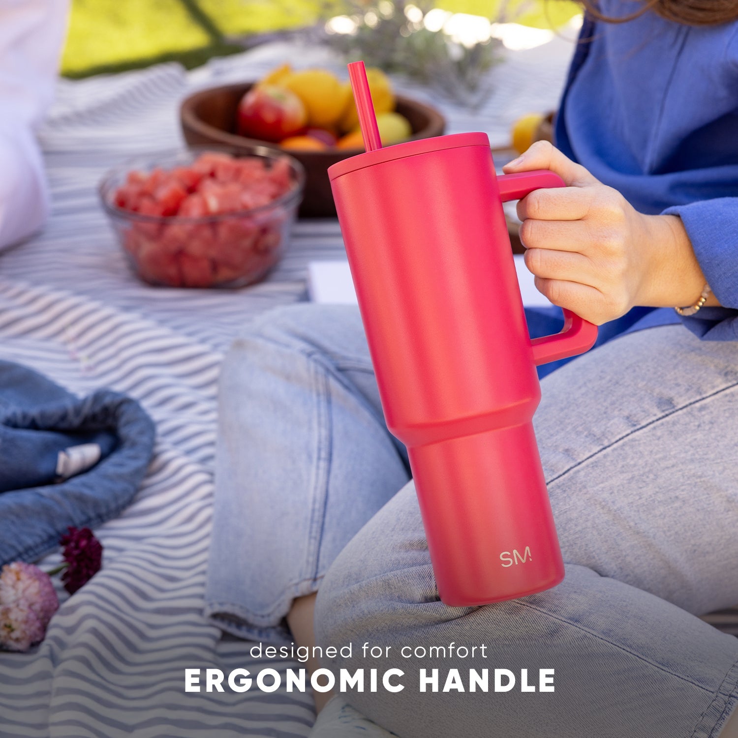 Coral pink insulated tumbler with ergonomic handle held by person in blue top during outdoor picnic, with fresh berries and fruits nearby on striped blanket. Text reads "designed for comfort" and "ERGONOMIC HANDLE."